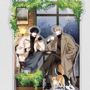 Peut inclure: Un présentoir en acrylique illustrant deux personnages de style anime assis à une table de café. L'un porte une veste crème et un jean foncé, l'autre un manteau beige et un pantalon sombre. Un chat roux et un chien noir et blanc sont à leurs pieds.