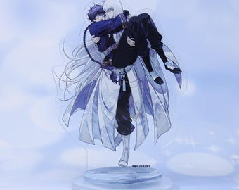 Op de afbeelding: Acrylstandaard met een anime-stijl illustratie van twee figuren die elkaar omarmen. Eén figuur met lang wit haar draagt een ander met donker haar. De standaard heeft een heldere basis en een lichtblauwe achtergrond.