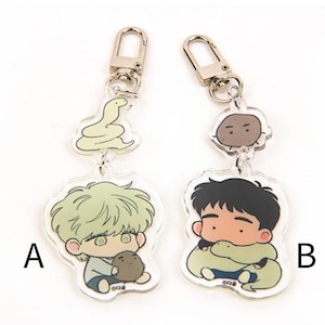 May include: Two clear acrylic keychains with cartoon characters. Keychain A features a green snake and a character with green hair holding a brown object. Keychain B features a brown character holding a green snake. Both keychains have silver metal rings.