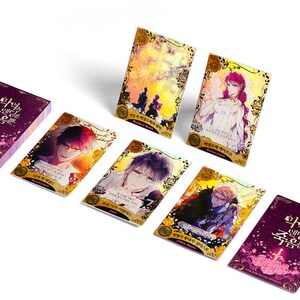 May include: A set of 7 collectible cards featuring characters from the graphic novel "The Holy Circle of the Sword". The cards are gold and purple with holographic accents. The cards feature different characters from the novel, including a woman in a white dress, a man with purple hair, and a man with black hair.