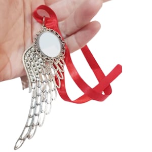 May include: A silver-colored angel wing ornament with a blank, oval-shaped center. The wing has detailed feather designs. A red ribbon is attached for hanging. The ornament is held in a hand against a white background.
