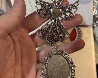 May include: A silver-toned metal angel ornament featuring an openwork filigree design on the wings and dress, a smooth heart-shaped centre, and a halo. An oval-shaped pendant frame with an ornate, scalloped border hangs from the base of the angel.