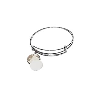 May include: A silver-colored bangle bracelet with two round charms. One charm has a white surface, and the other has a partially visible label. The bracelet is made of shiny metal and is set against a white background.