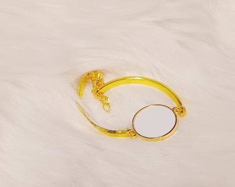 May include: A gold bracelet featuring a round, white, blank charm. The bracelet is made from a thin, gold chain and a thick, gold band.
