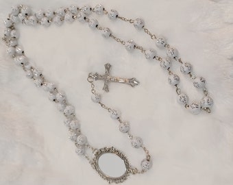 May include: White rosary beads with a silver cross and a silver oval mirror charm.