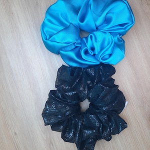 Peut inclure: Deux chouchous pour cheveux, l'un d'un bleu turquoise vif et uni avec une finition lisse et satinée, et l'autre d'un noir foncé avec un motif texturé subtil et répétitif rappelant une peau de serpent sur toute la surface du tissu.