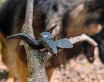 May include: A dark metal bird sculpture perched on a small tree branch. The sculpture has outstretched wings and is set against a blurred background of a German Shepherd dog and a forest setting.