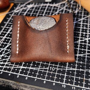 Op de afbeelding: Een bruine leren muntportemonnee met witte stiksels. De portemonnee heeft een klep die omslaat om de inhoud te beveiligen.