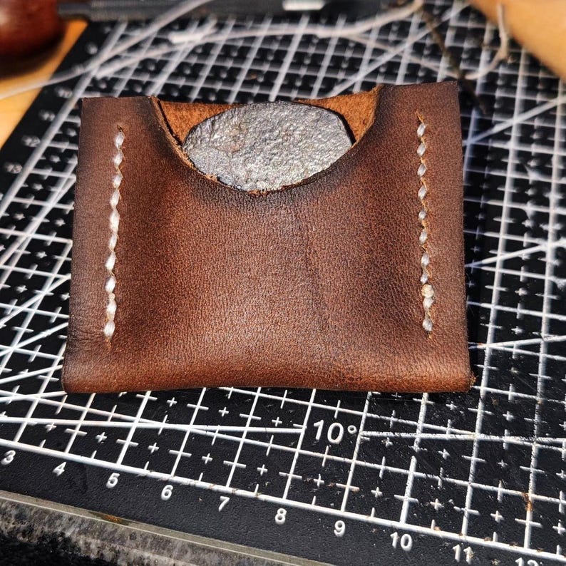 Op de afbeelding: Een bruine leren muntportemonnee met witte stiksels. De portemonnee heeft een klep die omslaat om de inhoud te beveiligen.