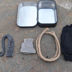 May include: A survival kit with a metal tin, a ferro rod, a flint striker, a piece of flint, a length of rope, and a piece of black cloth. The items are arranged on a large, gray rock.