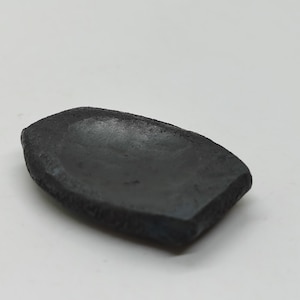 Op de afbeelding: Een klein, zwart, keramisch schaaltje met een licht afgeronde, ovale vorm. Het schaaltje heeft een ruw, getextureerd oppervlak en een licht ingedeukt midden.