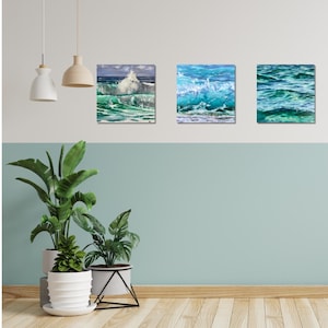 May include: Three framed paintings of ocean waves. The paintings are in shades of blue and green, with white foam crests. The paintings are hung on a white wall with a light blue accent wall below.