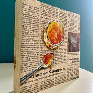 May include: A wooden block with a newspaper clipping about the reform of the power grid. The clipping is painted over with a spoon holding a dollop of orange and yellow paint. The text on the clipping reads "Reform des Stromsystems".