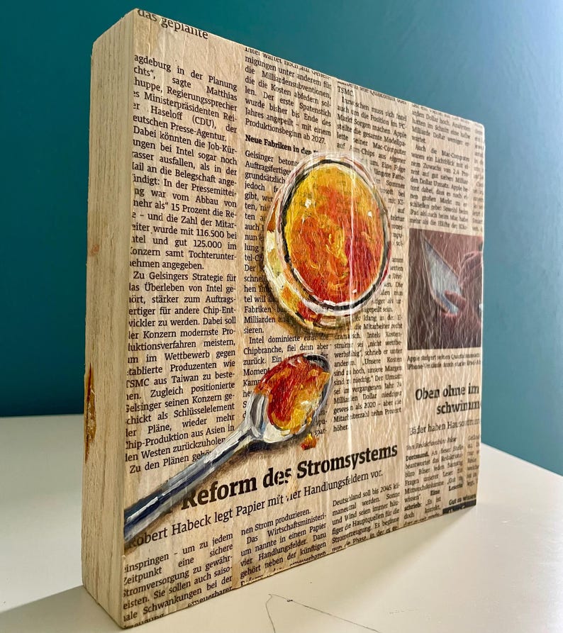 May include: A wooden block with a newspaper clipping about the reform of the power grid. The clipping is painted over with a spoon holding a dollop of orange and yellow paint. The text on the clipping reads "Reform des Stromsystems".