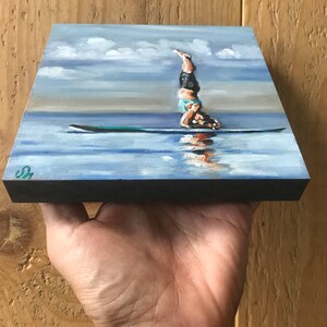 May include: A painting of a person doing a handstand on a paddleboard in the water. The sky is blue with white clouds. The water is a light blue color. The paddleboard is green.