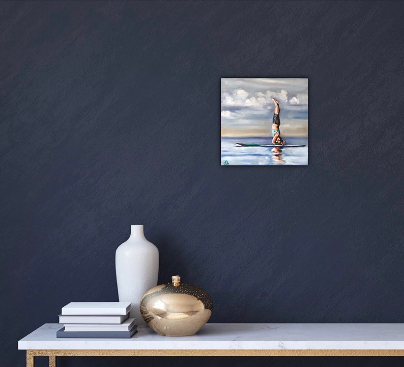 May include: A painting of a person doing a handstand on a paddleboard in the water. The sky is blue with white clouds. The water is a light blue color.