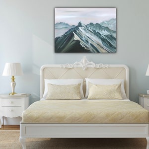 May include: A white bed frame with a beige headboard and a beige quilted bedspread. Two beige pillows with a geometric pattern are on the bed. A painting of a mountain range in shades of blue and green hangs above the bed.
