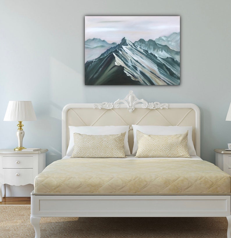 May include: A white bed frame with a beige headboard and a beige quilted bedspread. Two beige pillows with a geometric pattern are on the bed. A painting of a mountain range in shades of blue and green hangs above the bed.
