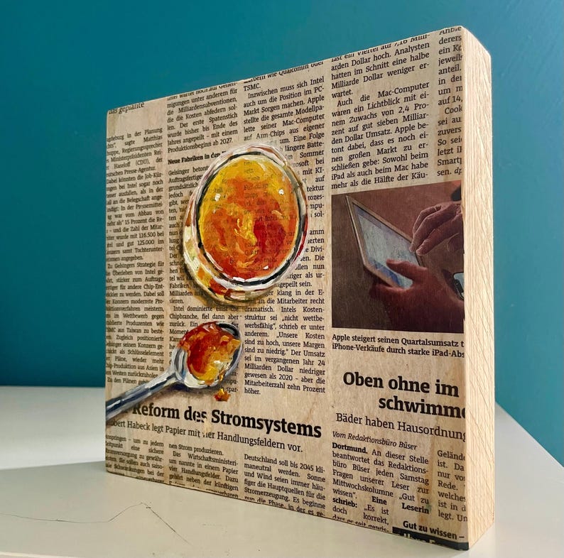 May include: A close-up of a wooden block with a collage of newspaper clippings and painted images. The image includes a glass of orange juice, a spoon with orange jam, and a hand holding a tablet. The text on the newspaper clippings includes the words "Reform des Stromsystems" and "Oben ohne im schwimmen".