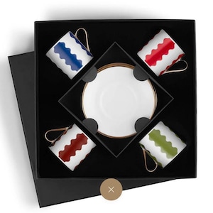 May include: A boxed set of four white espresso cups with wavy patterns in blue, red, and green, and a white saucer with a gold rim. The cups have gold handles and are arranged in a black box.