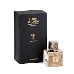 May include: A black box with gold lettering that reads "Parfum Artisanal Grande Pharmacie Parisienne Elixir 2 Extrait de Parfum Atelier Rebul" and a smaller bottle of perfume with the same text in gold lettering on a white background.