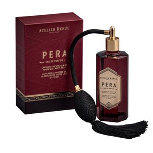 May include: A dark red box with gold lettering that reads "Atelier Rebul" and "Pera Eau de Parfum". The box is next to a dark red glass bottle with a gold cap and a black tassel. The bottle is labelled "Atelier Rebul Pera Eau de Parfum".