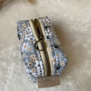 K&ouml;nnte beinhalten: Kleine, rechteckige Tasche mit floralem Muster, bronzefarbenem Rei&szlig;verschluss und beigem Zugband. Das Muster zeigt wei&szlig;e und beige Blumen auf hellblauem Hintergrund. Der Rei&szlig;verschluss ist ge&ouml;ffnet und gibt das beige Innere frei.