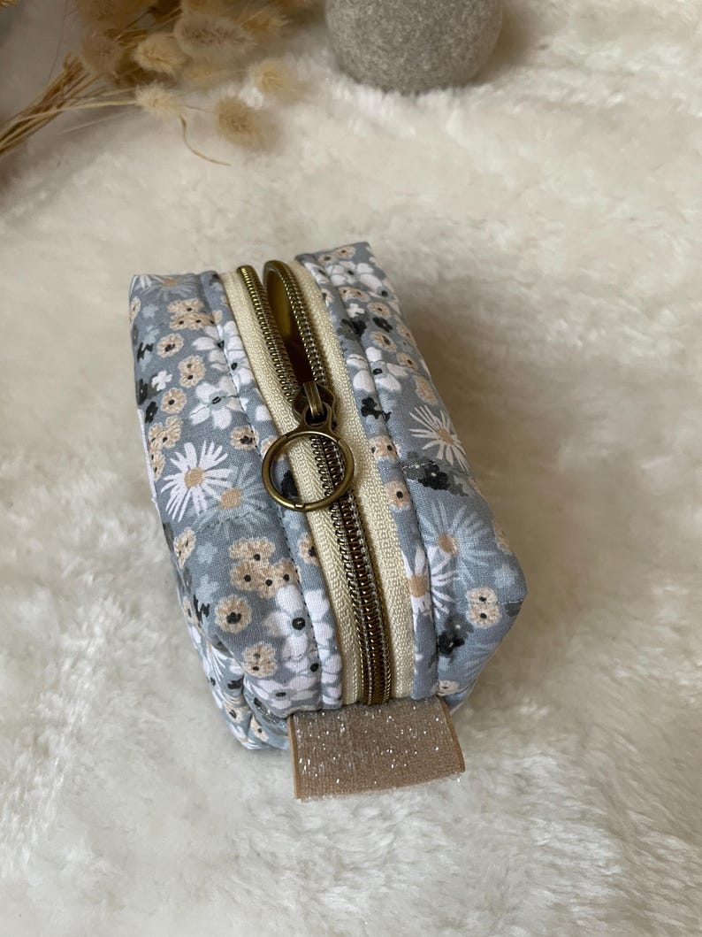 K&ouml;nnte beinhalten: Kleine, rechteckige Tasche mit floralem Muster, bronzefarbenem Rei&szlig;verschluss und beigem Zugband. Das Muster zeigt wei&szlig;e und beige Blumen auf hellblauem Hintergrund. Der Rei&szlig;verschluss ist ge&ouml;ffnet und gibt das beige Innere frei.