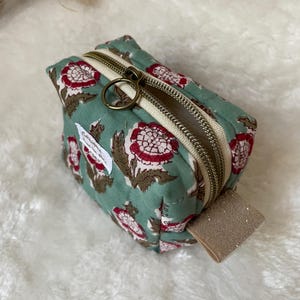 K&ouml;nnte beinhalten: Ein kleiner, quadratischer, t&uuml;rkisfarbener Beutel mit einem Blumenmuster in Rot und Braun. Der Beutel hat einen beigen Rei&szlig;verschluss und einen goldfarbenen Rei&szlig;verschlussanh&auml;nger. Ein kleines beiges Etikett ist an der Seite befestigt.