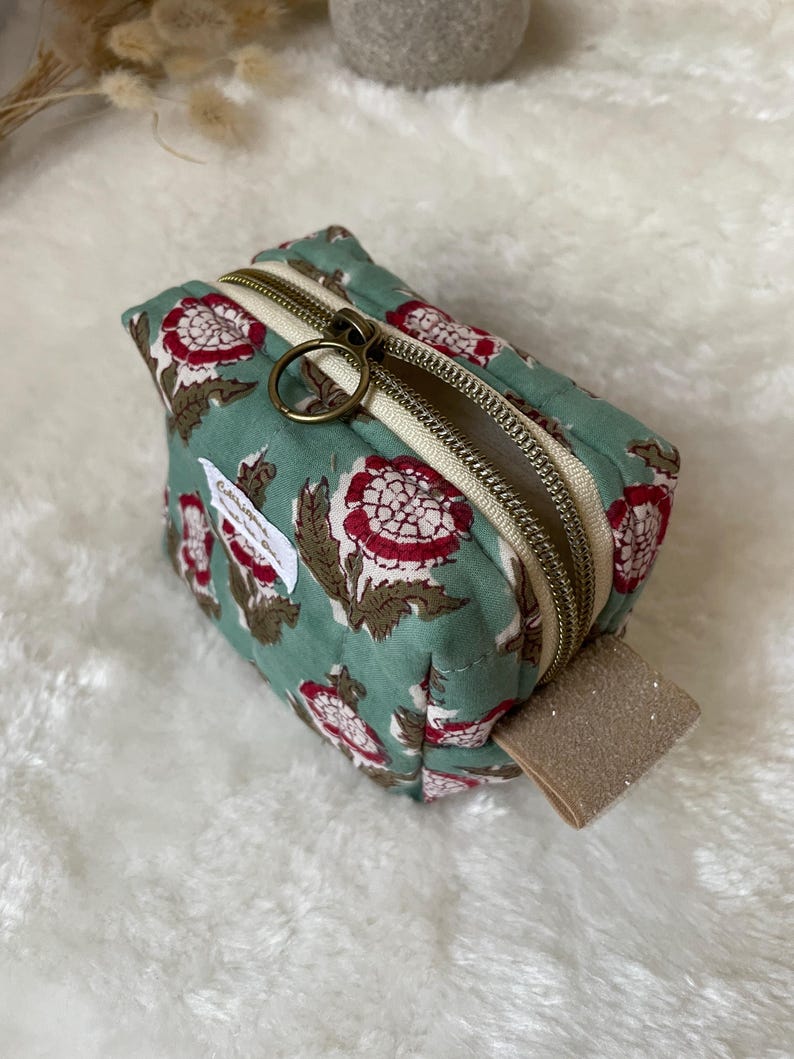 K&ouml;nnte beinhalten: Ein kleiner, quadratischer, t&uuml;rkisfarbener Beutel mit einem Blumenmuster in Rot und Braun. Der Beutel hat einen beigen Rei&szlig;verschluss und einen goldfarbenen Rei&szlig;verschlussanh&auml;nger. Ein kleines beiges Etikett ist an der Seite befestigt.