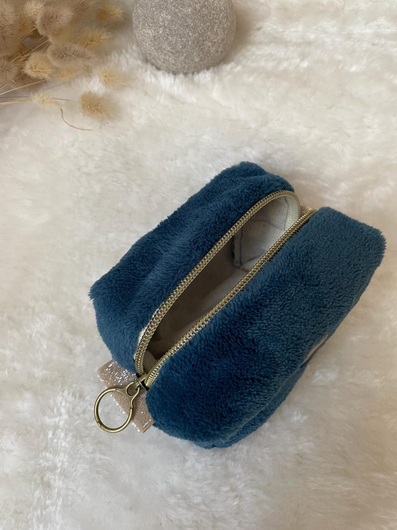K&ouml;nnte beinhalten: Ein rechteckiges, tiefblaues, pl&uuml;schiges Etui mit Rei&szlig;verschluss, goldfarbenem Rei&szlig;verschluss und einer kleinen, glitzernden Schleife. Das Etui ist ge&ouml;ffnet und zeigt ein helles Innenfutter und einen goldfarbenen Ring am Rei&szlig;verschluss.
