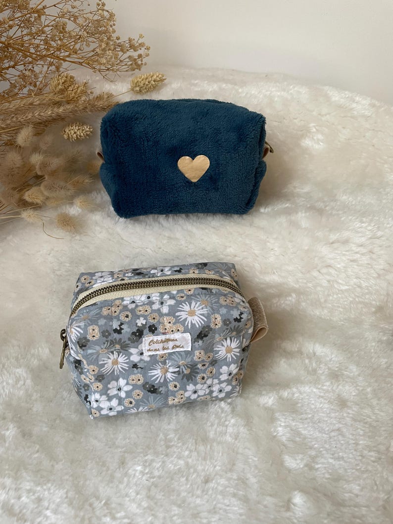 K&ouml;nnte beinhalten: Zwei kleine Kosmetiktaschen. Die obere Tasche ist aus tiefblauem Pl&uuml;sch mit einem Holzherz-Akzent. Die untere Tasche ist grau mit einem Blumenmuster, einem Rei&szlig;verschluss und einem kleinen Etikett "Colchiques dans les pr&eacute;s."