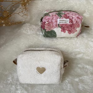 K&ouml;nnte beinhalten: Zwei kleine Kosmetiktaschen. Eine ist wei&szlig; mit einem goldenen Herz, die andere ist floral mit rosa und wei&szlig;en Hortensienbl&uuml;ten. Beide haben Rei&szlig;verschl&uuml;sse und kleine braune Lederlaschen.