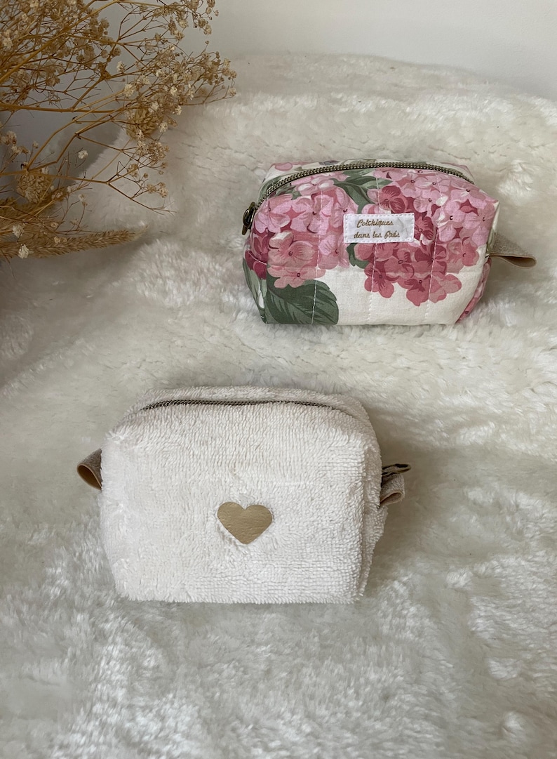 K&ouml;nnte beinhalten: Zwei kleine Kosmetiktaschen. Eine ist wei&szlig; mit einem goldenen Herz, die andere ist floral mit rosa und wei&szlig;en Hortensienbl&uuml;ten. Beide haben Rei&szlig;verschl&uuml;sse und kleine braune Lederlaschen.