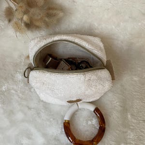 K&ouml;nnte beinhalten: Eine wei&szlig;e, flauschige Rei&szlig;verschlusstasche mit goldenem Rei&szlig;verschluss, ge&ouml;ffnet, um eine Uhr und Schmuck zu zeigen. Ein wei&szlig;es und braunes Armband liegt vor der Tasche. Getrocknete Blumen sind im Hintergrund.