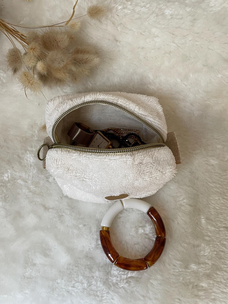 K&ouml;nnte beinhalten: Eine wei&szlig;e, flauschige Rei&szlig;verschlusstasche mit goldenem Rei&szlig;verschluss, ge&ouml;ffnet, um eine Uhr und Schmuck zu zeigen. Ein wei&szlig;es und braunes Armband liegt vor der Tasche. Getrocknete Blumen sind im Hintergrund.