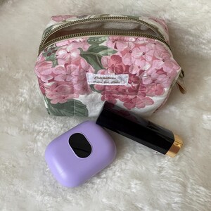 K&ouml;nnte beinhalten: Eine Kosmetiktasche mit floralem Muster mit Rei&szlig;verschluss, einem lila Etui und einer schwarzen Lippenstift-Tube mit goldenem Boden. Die Tasche zeigt rosa Bl&uuml;ten und gr&uuml;ne Bl&auml;tter auf wei&szlig;em Grund. Die Tasche tr&auml;gt den Text "Colchiques dans ton pr&eacute;s".