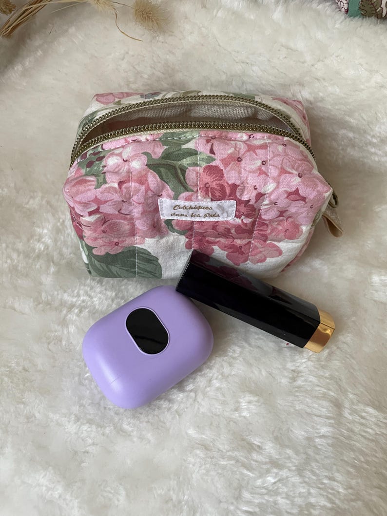 K&ouml;nnte beinhalten: Eine Kosmetiktasche mit floralem Muster mit Rei&szlig;verschluss, einem lila Etui und einer schwarzen Lippenstift-Tube mit goldenem Boden. Die Tasche zeigt rosa Bl&uuml;ten und gr&uuml;ne Bl&auml;tter auf wei&szlig;em Grund. Die Tasche tr&auml;gt den Text "Colchiques dans ton pr&eacute;s".