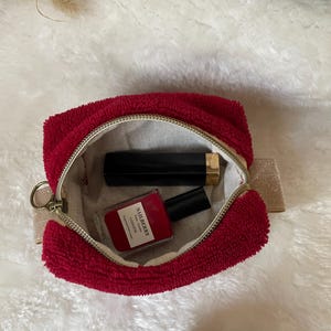 K&ouml;nnte beinhalten: Eine rote Kosmetiktasche aus Frottee mit goldenem Rei&szlig;verschluss, ge&ouml;ffnet, um eine schwarze Lippenstift-Tube und eine Flasche roten Nagellacks zu zeigen. Auf der Nagellackflasche steht "NAILBERRY". Die Tasche hat eine kleine goldene Schlaufe.