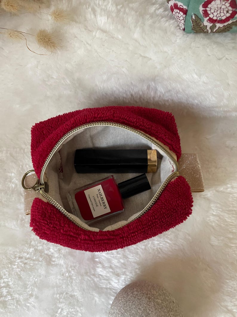 K&ouml;nnte beinhalten: Eine rote Kosmetiktasche aus Frottee mit goldenem Rei&szlig;verschluss, ge&ouml;ffnet, um eine schwarze Lippenstift-Tube und eine Flasche roten Nagellacks zu zeigen. Auf der Nagellackflasche steht "NAILBERRY". Die Tasche hat eine kleine goldene Schlaufe.