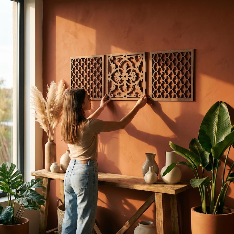 May include: Three wooden wall panels with intricate geometric and floral cut-out designs are being hung on a terracotta-colored wall. The panels are rectangular and have a natural wood finish. The scene is set in a well-lit room with a rustic wooden console table.