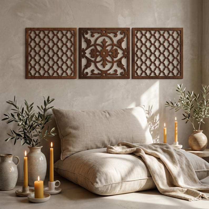 May include: Three square, wooden wall panels with intricate, openwork designs in a warm brown hue. Below, a large, neutral-toned pillow and throw are arranged, with lit candles and vases of greenery adding to the cozy ambiance.