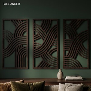 May include: Three dark brown wooden wall panels with a wave-like design against a dark green wall. The panels are rectangular with a repeating pattern of curved lines. A small vase and books are on a wooden surface below.