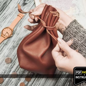 May include: A brown leather pouch with a drawstring closure. The pouch is being held by a person's hands. The text "PDF PATTERN + INSTRUCTION" is visible in the lower right corner of the image.