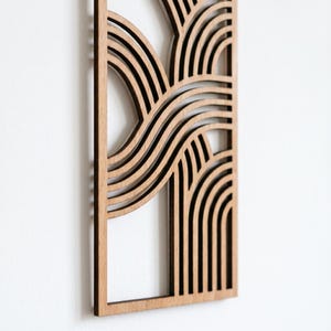 May include: A wooden wall art piece with a geometric design. The art features layered, curved lines and arches, creating a wave-like pattern. The wood has a natural, light brown color, and the piece is framed.