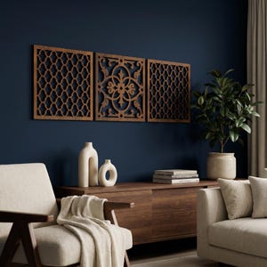 May include: Three wooden wall panels with intricate cutout designs are displayed against a dark blue wall. The panels are rectangular and feature geometric and floral patterns. A wooden cabinet, vases, and a plant in a pot are visible below the panels.