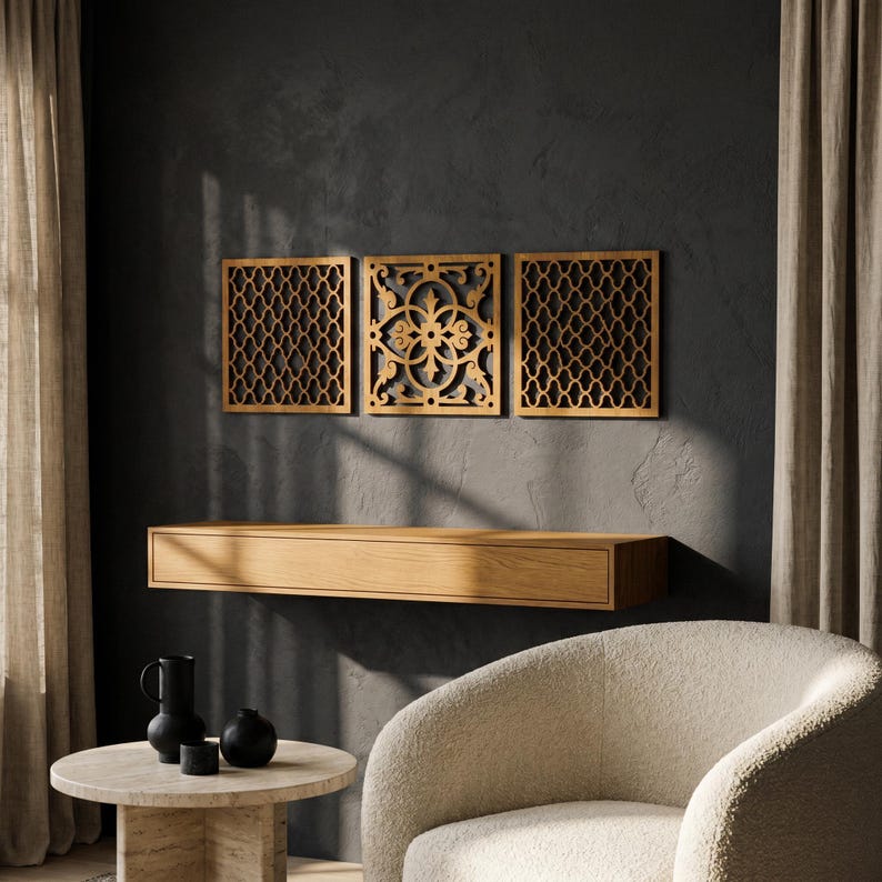 May include: Three wooden wall art panels with intricate geometric and floral cut-out designs. The panels are mounted above a floating wooden shelf. The room features a neutral color palette with a dark gray wall, beige curtains, and a cream-colored armchair.