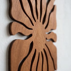 K&ouml;nnte beinhalten: Holzwandkunst mit einem stilisierten Blattdesign. Das Kunstwerk besteht aus hellbraunem Holz mit ausgeschnittenen Details, wodurch ein geschichteter Effekt entsteht. Es ist an einer wei&szlig;en Wand befestigt und zeigt die nat&uuml;rliche Holzmaserung.