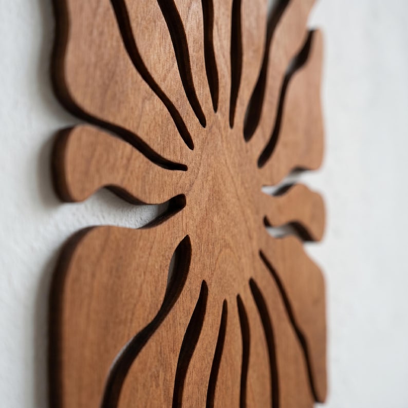 K&ouml;nnte beinhalten: Holzwandkunst mit einem stilisierten Blattdesign. Das Kunstwerk besteht aus hellbraunem Holz mit ausgeschnittenen Details, wodurch ein geschichteter Effekt entsteht. Es ist an einer wei&szlig;en Wand befestigt und zeigt die nat&uuml;rliche Holzmaserung.