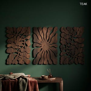 K&ouml;nnte beinhalten: Drei dunkelbraune Holz-Wandpaneele mit floralem Design sind an einer dunkelgr&uuml;nen Wand angebracht. Die Paneele sind aus Teakholz gefertigt. Ein Holztisch mit Schale, Tablett und Diffusor befindet sich im Vordergrund.